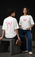 Two models wearing white graphic t-shirts with pink bow designs, one seated on a dark bench showing the back graphic and the other standing showing the front graphic, in a dark studio setting.